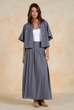 Load image into Gallery viewer, A woman models a matching two-piece pinstripe set consisting of a cropped short-sleeve button-up shirt and a high-waisted pleated maxi skirt, styled over a white tank top and beige boots against a textured beige backdrop.
