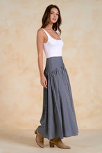 Load image into Gallery viewer, A woman modeling a white ribbed tank top paired with a high-waisted, floor-length pleated skirt in navy and white vertical stripes, styled with brown suede boots against a textured beige backdrop.

