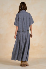 Load image into Gallery viewer, A woman shown from the back wearing a matching short-sleeve collared shirt and pleated maxi skirt set in a blue and white vertical stripe pattern, set against a warm beige textured studio backdrop.
