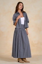 Load image into Gallery viewer, A woman models a matching navy and white vertically striped shirt jacket and midi skirt set, styled with a white tank top and beige ankle boots against a textured beige backdrop.

