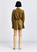 Load image into Gallery viewer, A model viewed from the back wearing an olive green long-sleeve collared shirt and matching mini skirt with a tie belt, paired with beige knee-high boots, against a light gray studio background.
