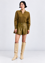 Load image into Gallery viewer, A model stands against a light gray studio background wearing a matching olive green collared shirt and faux-leather mini skirt, paired with beige knee-high boots.
