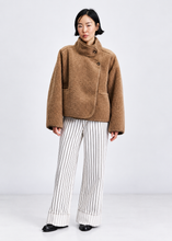 Load image into Gallery viewer, A model stands against a light gray studio background wearing a brown bouclé cropped jacket with a high mock neck and front buttons, paired with white wide-leg trousers featuring thin black vertical stripes, and black shoes.
