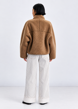 Load image into Gallery viewer, A model viewed from the back wearing a camel brown fuzzy cropped jacket and white wide-leg trousers with thin black vertical stripes, set against a plain light gray studio background.
