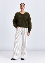 Load image into Gallery viewer, A model stands against a light gray studio background wearing a fuzzy olive green crewneck sweater and white wide-leg trousers with thin black vertical stripes.
