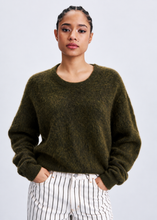 Load image into Gallery viewer, A woman wearing a fuzzy, textured olive green crewneck sweater and white pants with thin black vertical stripes, standing against a light gray studio background.

