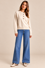Load image into Gallery viewer, A woman modeling a cream knit sweater with brown buttons and wide-leg blue jeans, standing against a solid beige studio background.
