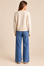 Load image into Gallery viewer, A woman shown from the back wearing a cream knit sweater and wide-leg blue jeans, standing against a solid beige studio background.
