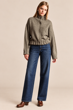 Load image into Gallery viewer, A model wearing a textured taupe mock-neck knit sweater and dark blue wide-leg jeans, standing against a solid beige studio background.
