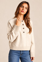 Load image into Gallery viewer, A woman wearing a cream-colored knit henley sweater with brown buttons and blue jeans, posing against a solid beige studio background.
